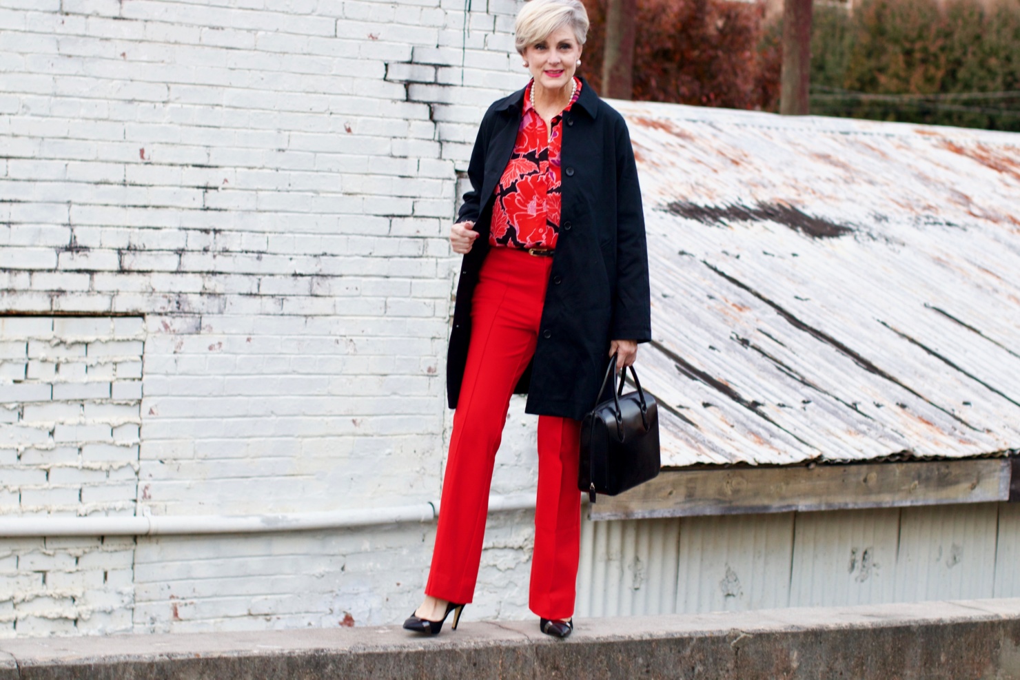 beth from Style at a Certain Age wears Anthropologie red pants, pink and red shirt, Everlane black raincoat, and black suede pumps