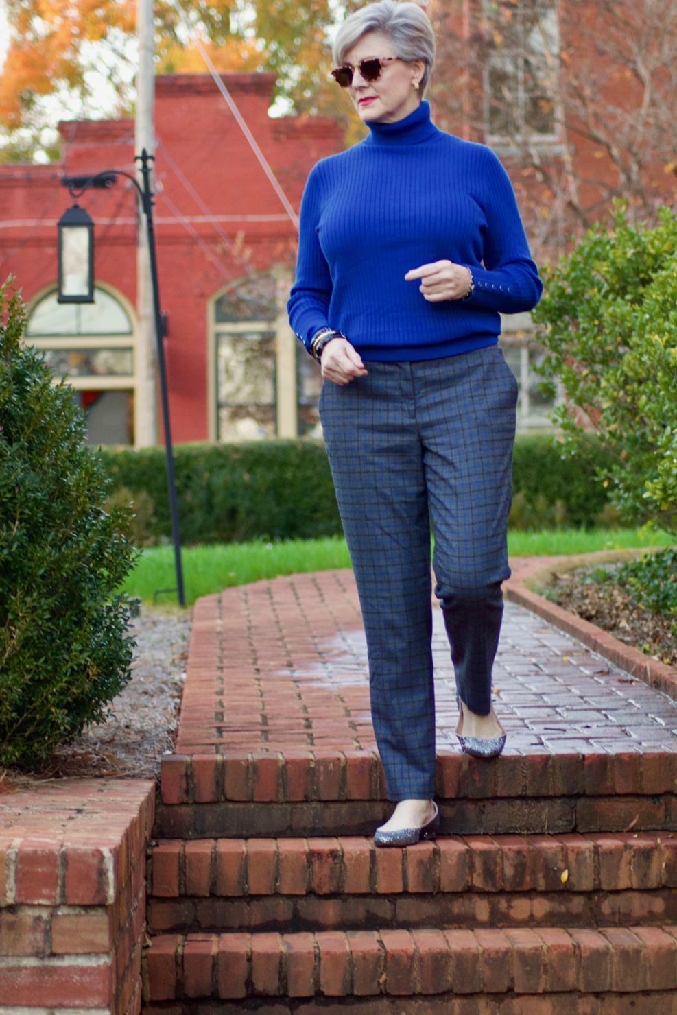 beth from style at a certain age wears plaid pants, cobalt blue turtleneck, green wool coat, blackwatch plaid cashmere scarf, and green beanie