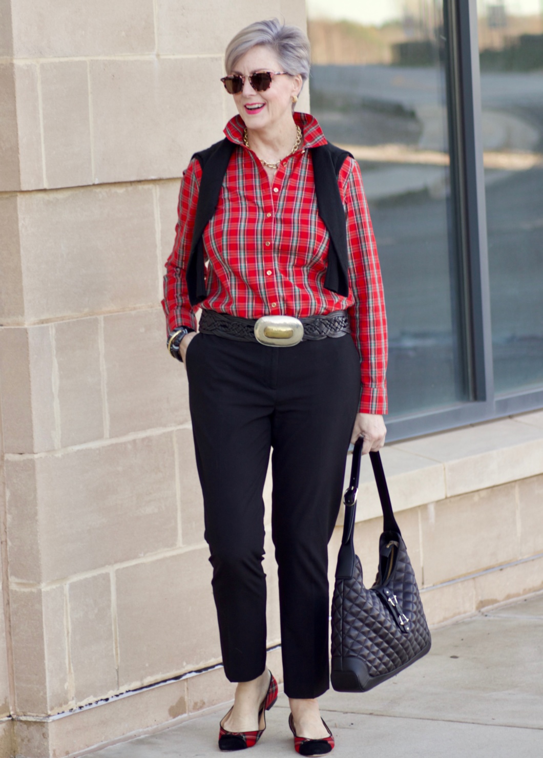 beth from Style at a Certain Age wears a tartan plaid shirt, black cropped ankle pants, tartan plaid flats, military coat and quilted hobo handbag.