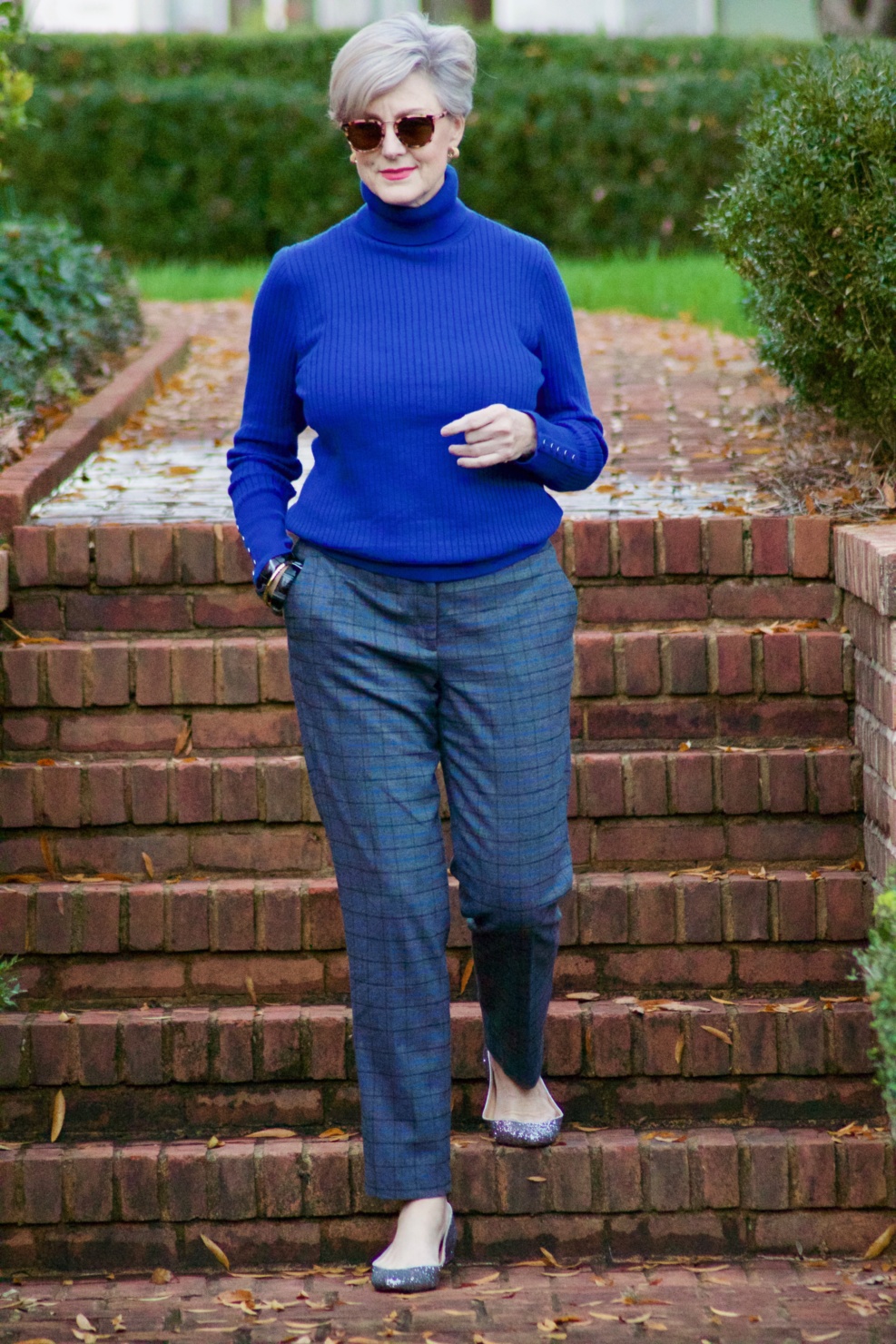 beth from style at a certain age wears plaid pants, cobalt blue turtleneck, green wool coat, blackwatch plaid cashmere scarf, and green beanie