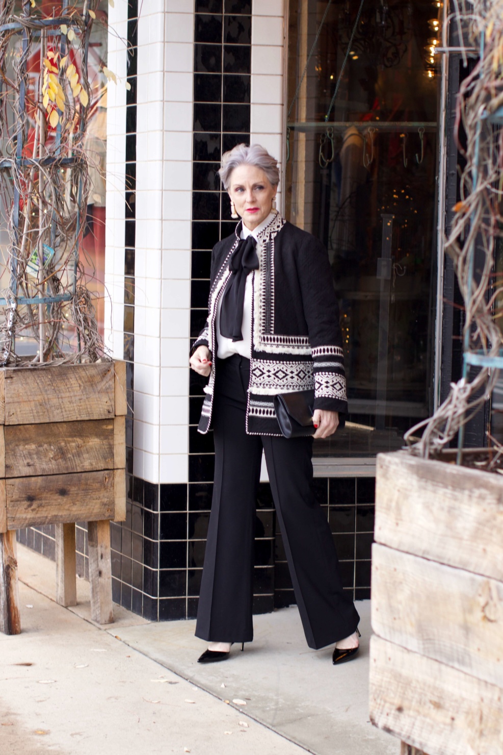 beth from Style at a Certain Age wears black flare pants, bow blouse, embroidered wonderland jacket, and black patent leather pumps