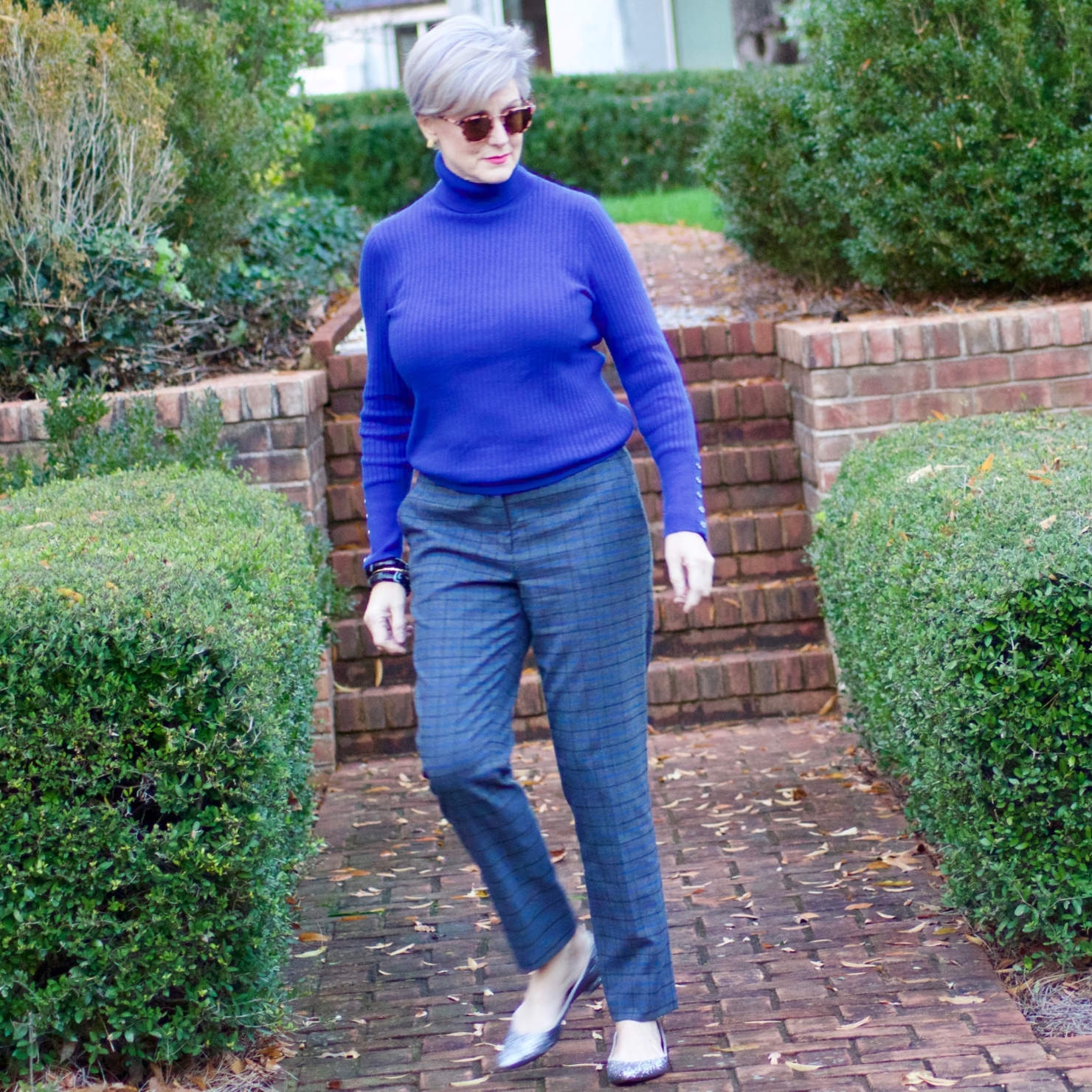 beth from style at a certain age wears plaid pants, cobalt blue turtleneck, green wool coat, blackwatch plaid cashmere scarf, and green beanie