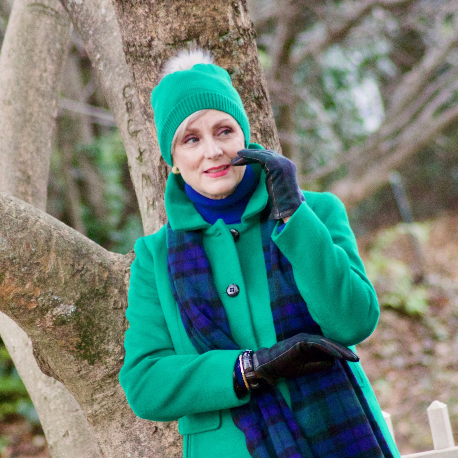 beth from style at a certain age wears plaid pants, cobalt blue turtleneck, green wool coat, blackwatch plaid cashmere scarf, and green beanie