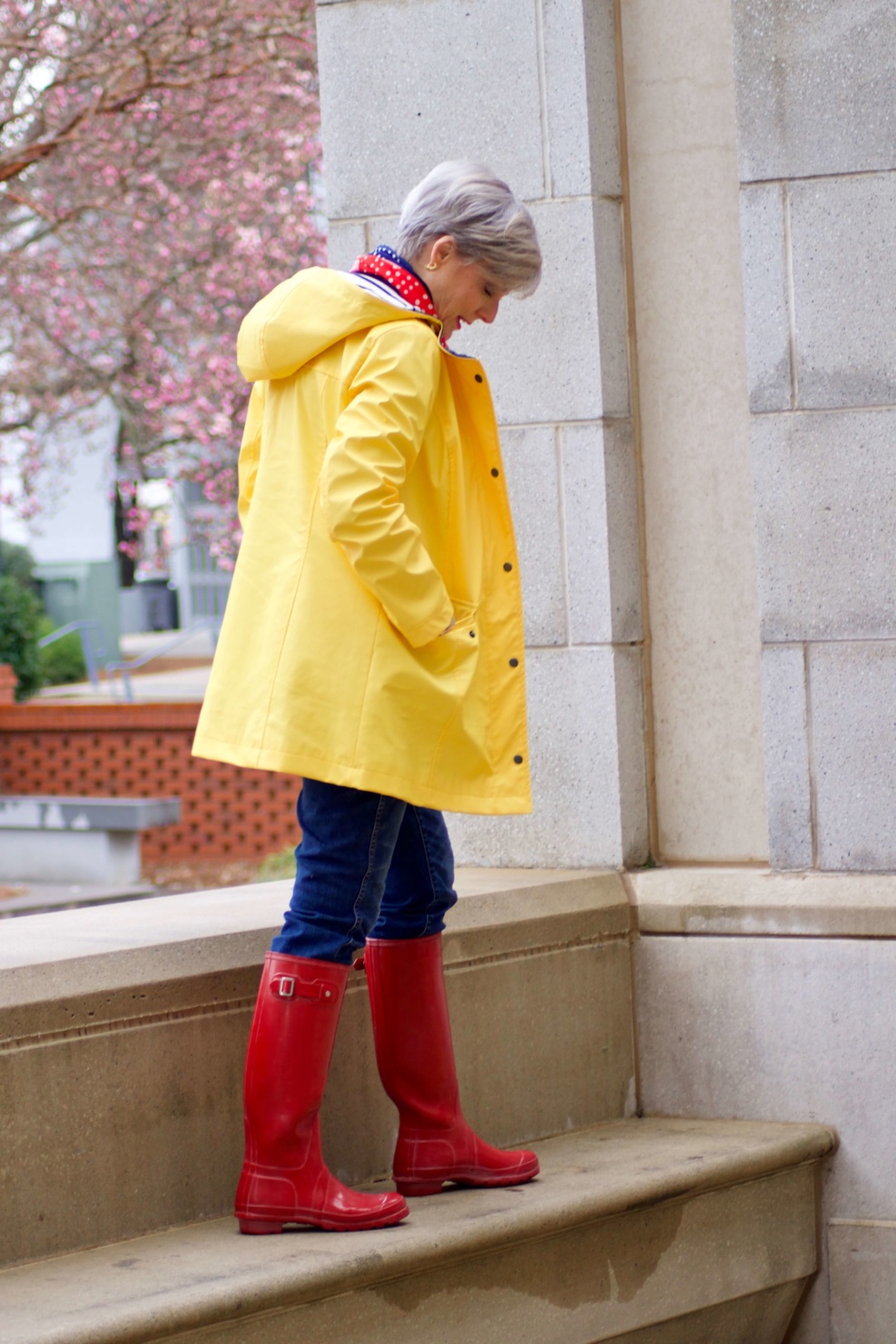 beth from Style at a Certain Age wears Hunter high gloss red rain boots, navy turtleneck, vintage straight jeans, yellow raincoat, and polka dot scarf