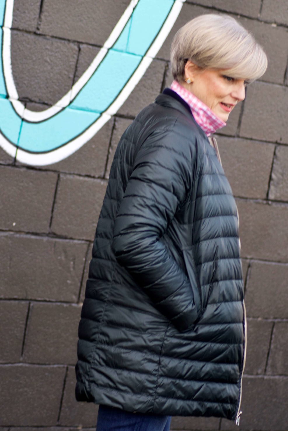 beth from Style at a Certain Age wears a puffer jacket, navy blue knit blazer, novelty sweater, pink gingham check shirt and blue jeans