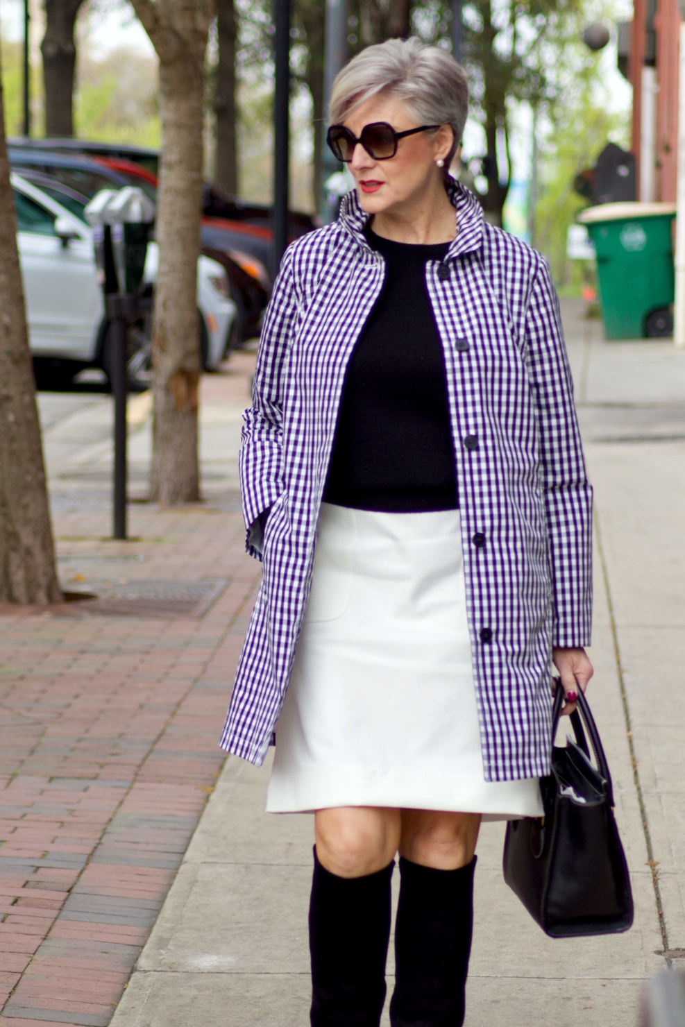 beth from Style at a Certain Age wears a gingham raincoat, white skirt, black sweater, and black boots