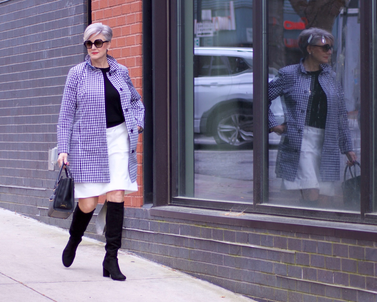 beth from Style at a Certain Age wears a gingham raincoat, white skirt, black sweater, and black boots