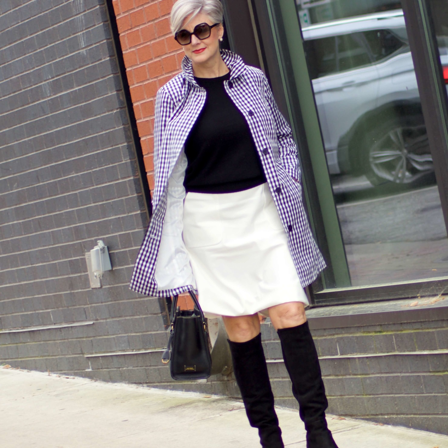 beth from Style at a Certain Age wears a gingham raincoat, white skirt, black sweater, and black boots