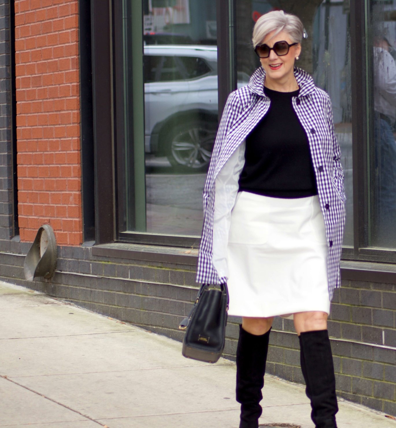 beth from Style at a Certain Age wears a gingham raincoat, white skirt, black sweater, and black boots