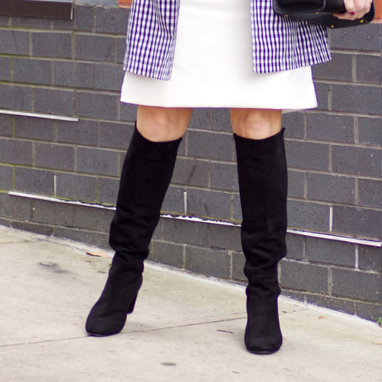 beth from Style at a Certain Age wears a gingham raincoat, white skirt, black sweater, and black boots