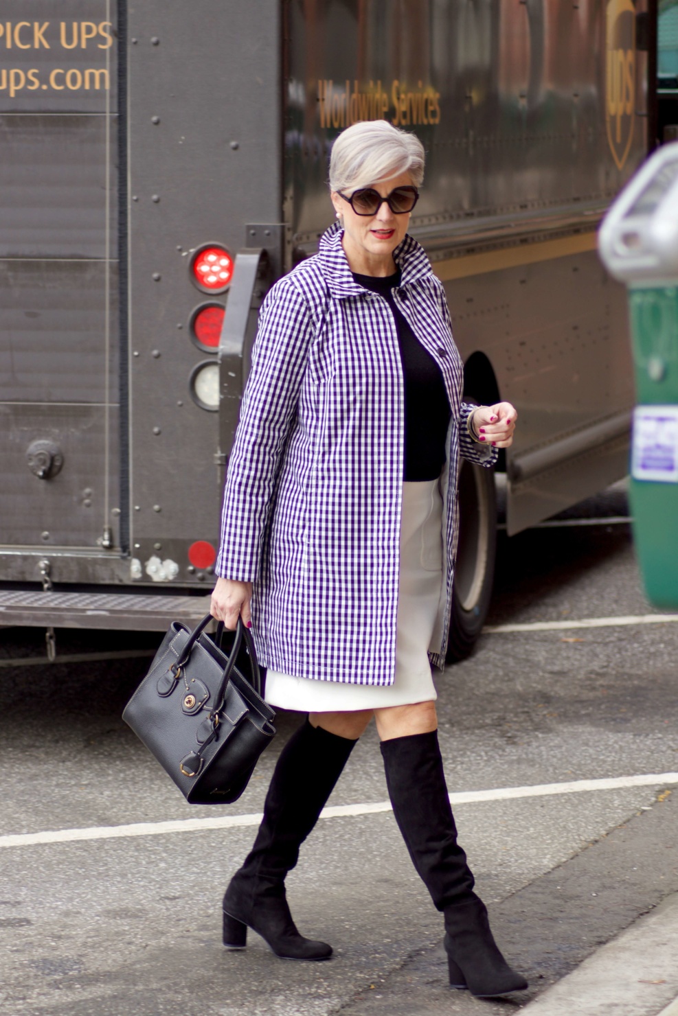 beth from Style at a Certain Age wears a gingham raincoat, white skirt, black sweater, and black boots