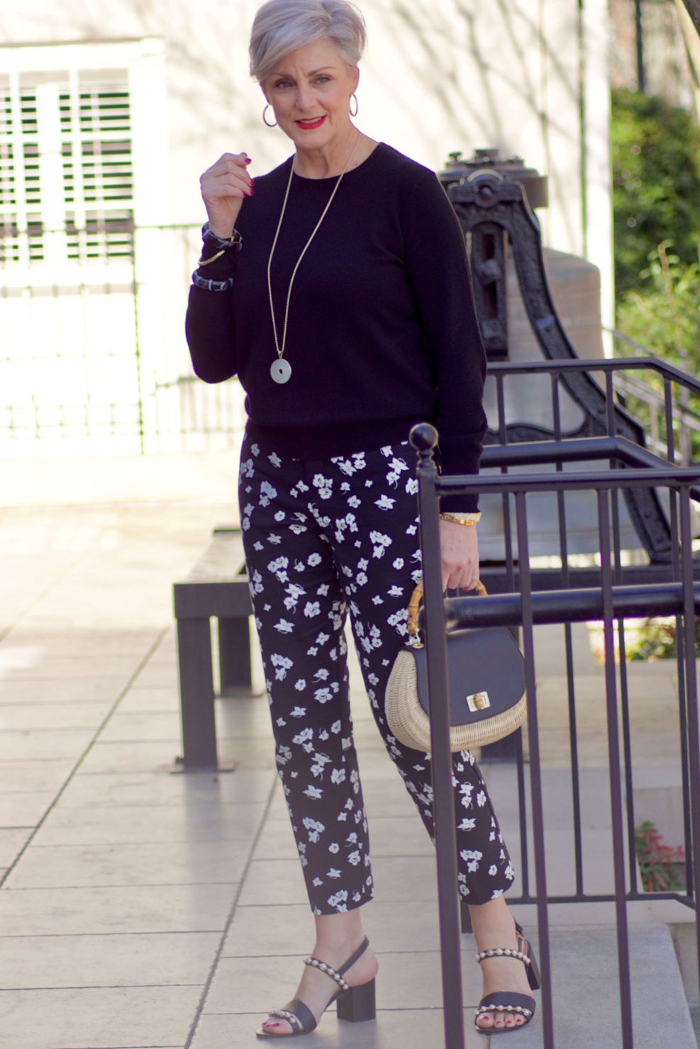 beth from Style at a Certain Age wears black and white floral ankle pants, black crewneck sweater, black pearl embellished sandals, and straw handbag