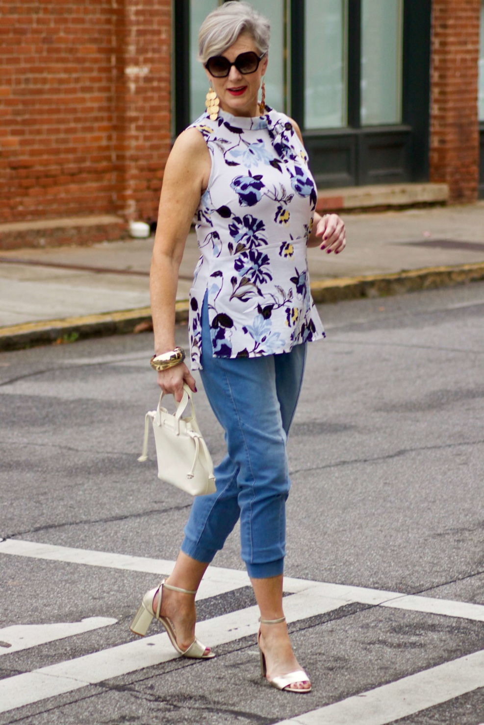 beth from Style at a Certain Age wears a floral top, denim joggers, gold metallic block sandals, and carries a drawstring crossbody handbag. Walmart we dress America