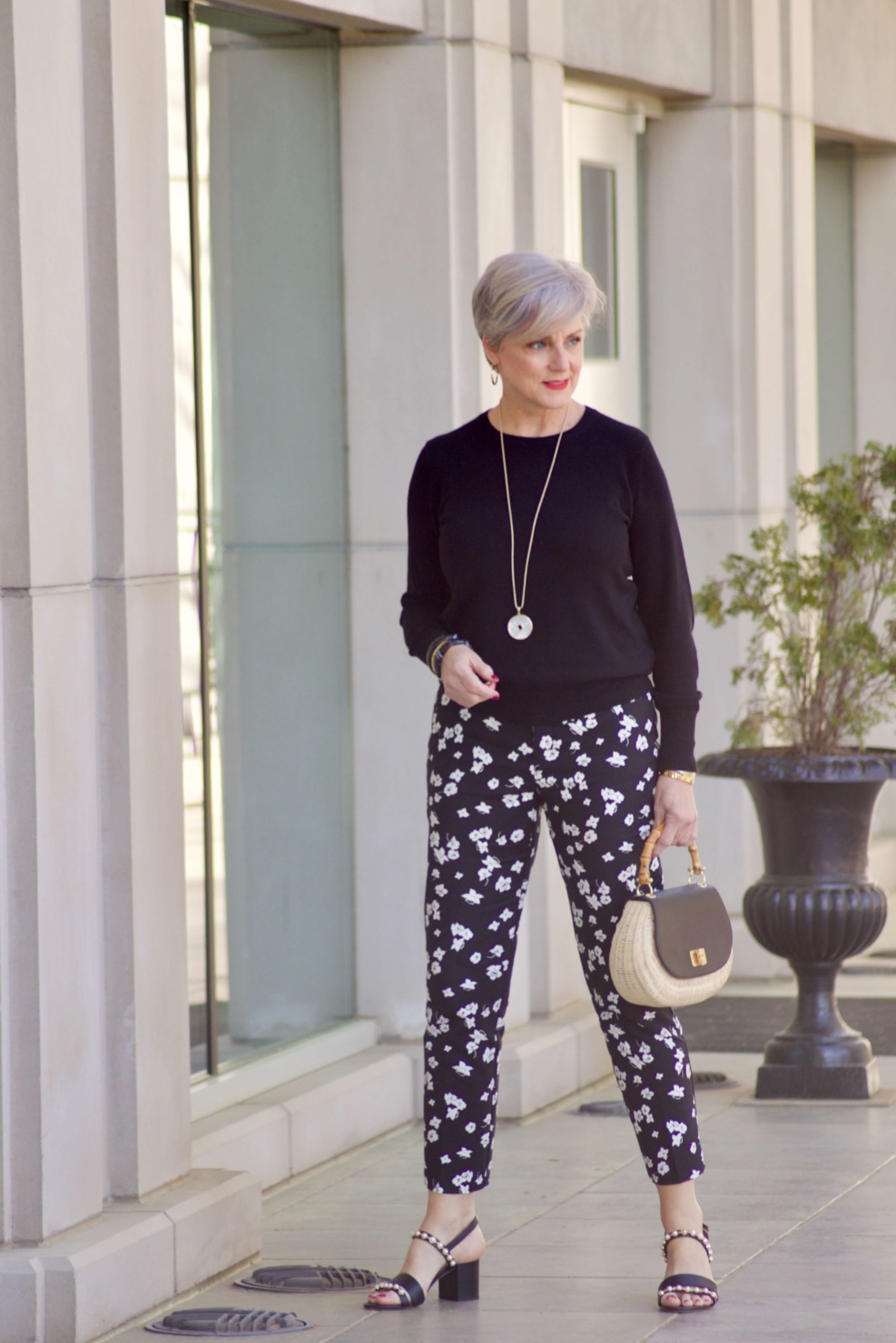 beth from Style at a Certain Age wears black and white floral ankle pants, black crewneck sweater, black pearl embellished sandals, and straw handbag