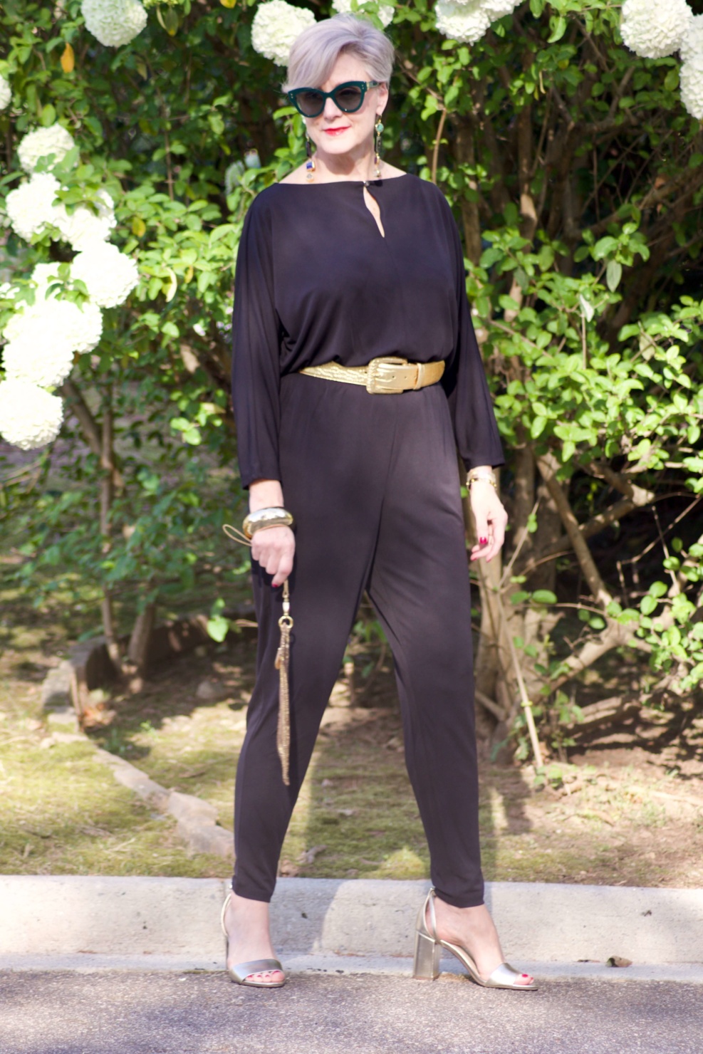 beth from Style at a Certain Age wears a black jumpsuit, gold sandals, gold belt, and gold clutch handbag