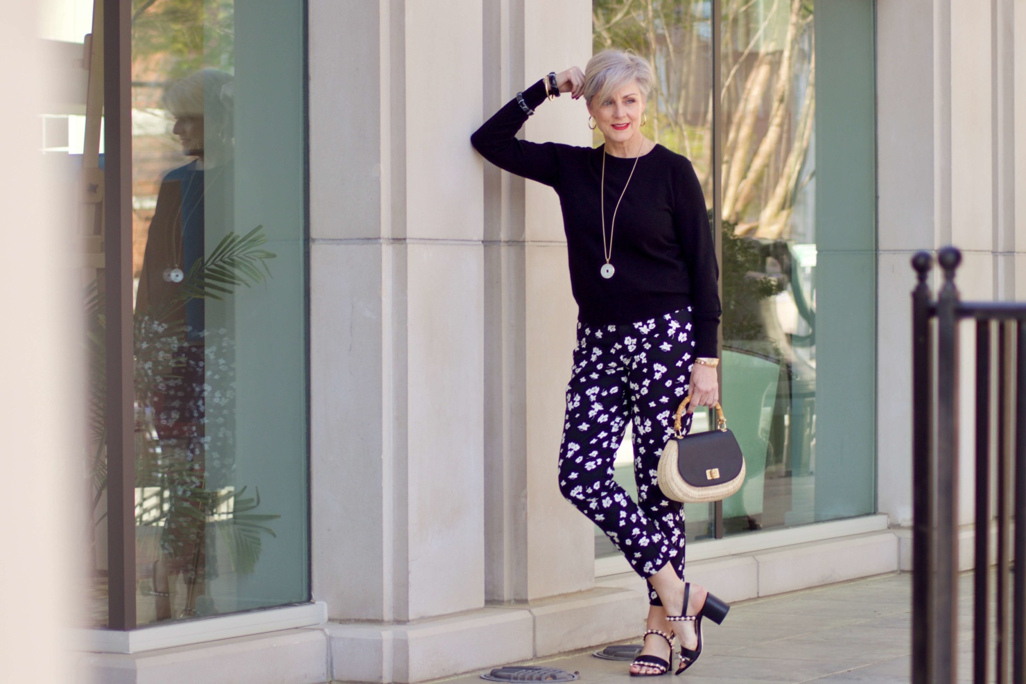 beth from Style at a Certain Age wears black and white floral ankle pants, black crewneck sweater, black pearl embellished sandals, and straw handbag