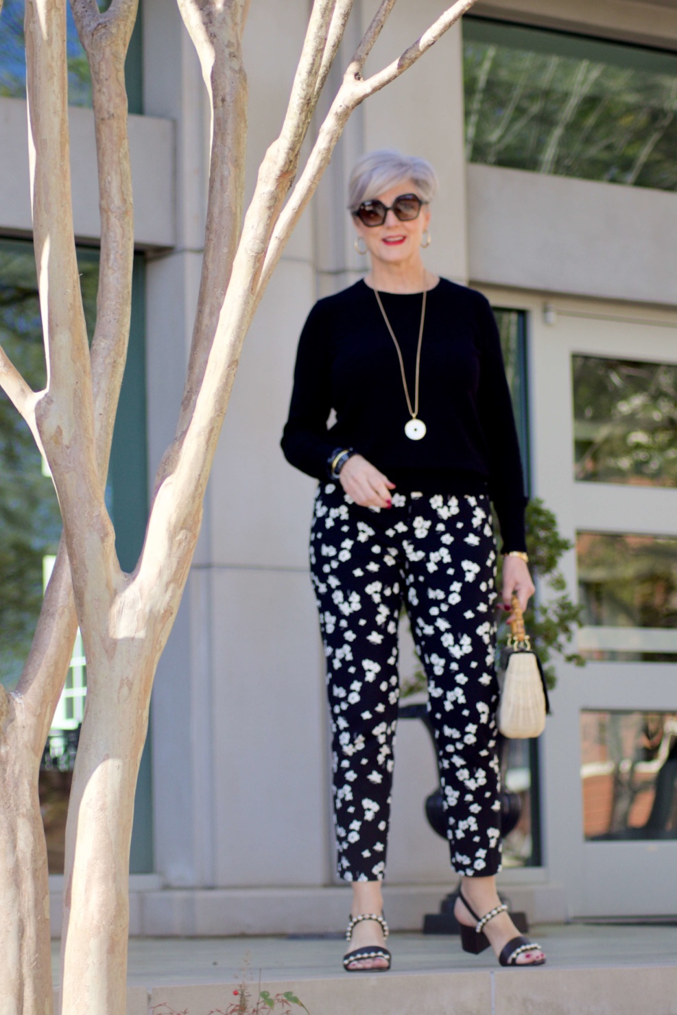 beth from Style at a Certain Age wears black and white floral ankle pants, black crewneck sweater, black pearl embellished sandals, and straw handbag