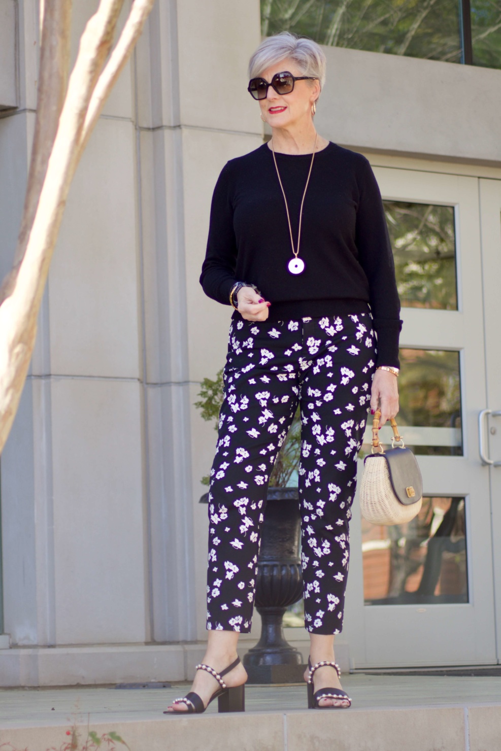 beth from Style at a Certain Age wears black and white floral ankle pants, black crewneck sweater, black pearl embellished sandals, and straw handbag
