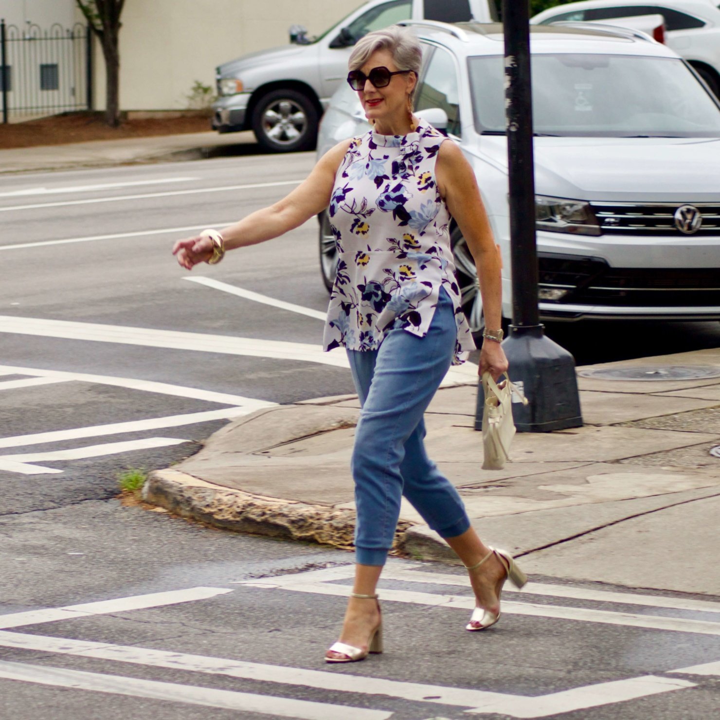 beth from Style at a Certain Age wears a floral top, denim joggers, gold metallic block sandals, and carries a drawstring crossbody handbag. Walmart we dress America