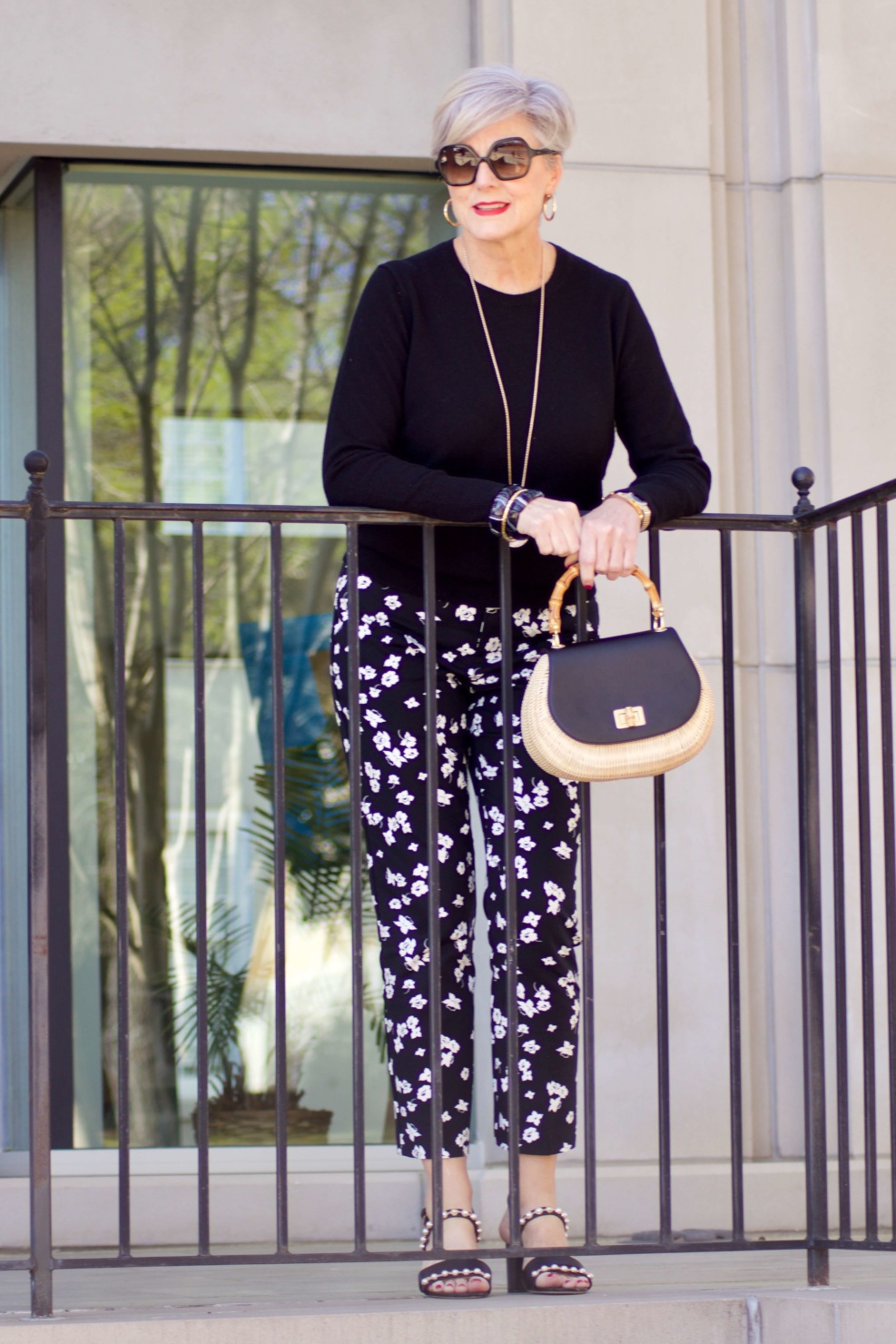 beth from Style at a Certain Age wears black and white floral ankle pants, black crewneck sweater, black pearl embellished sandals, and straw handbag