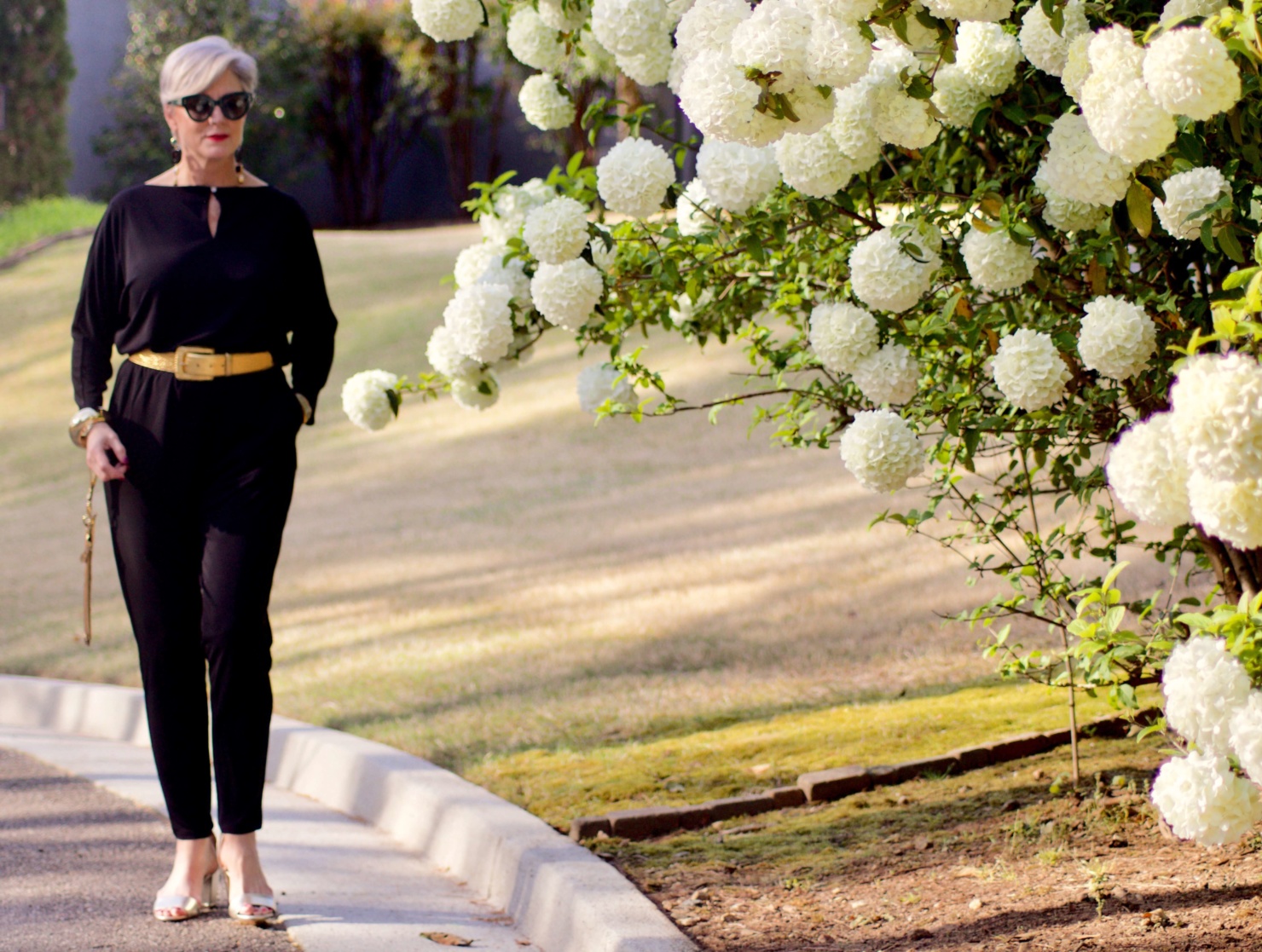 beth from Style at a Certain Age wears a black jumpsuit, gold sandals, gold belt, and gold clutch handbag