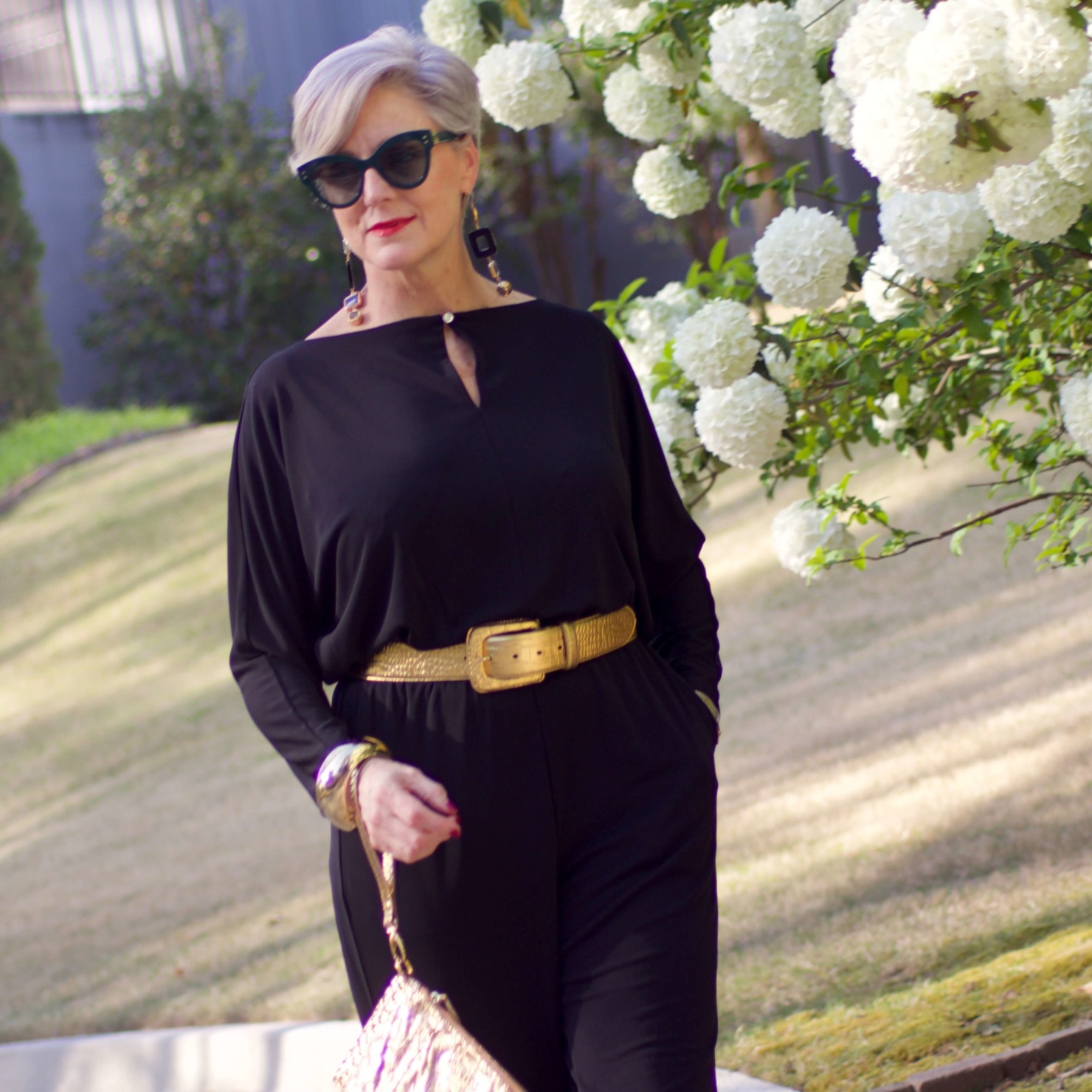 beth from Style at a Certain Age wears a black jumpsuit, gold sandals, gold belt, and gold clutch handbag