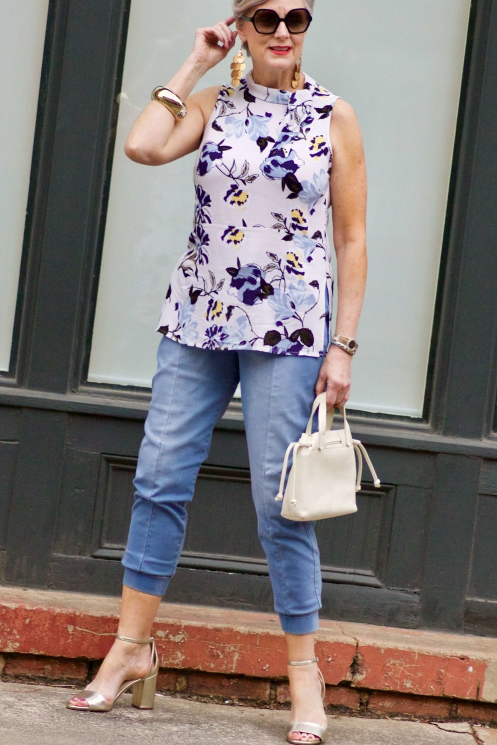 beth from Style at a Certain Age wears a floral top, denim joggers, gold metallic block sandals, and carries a drawstring crossbody handbag. Walmart we dress America