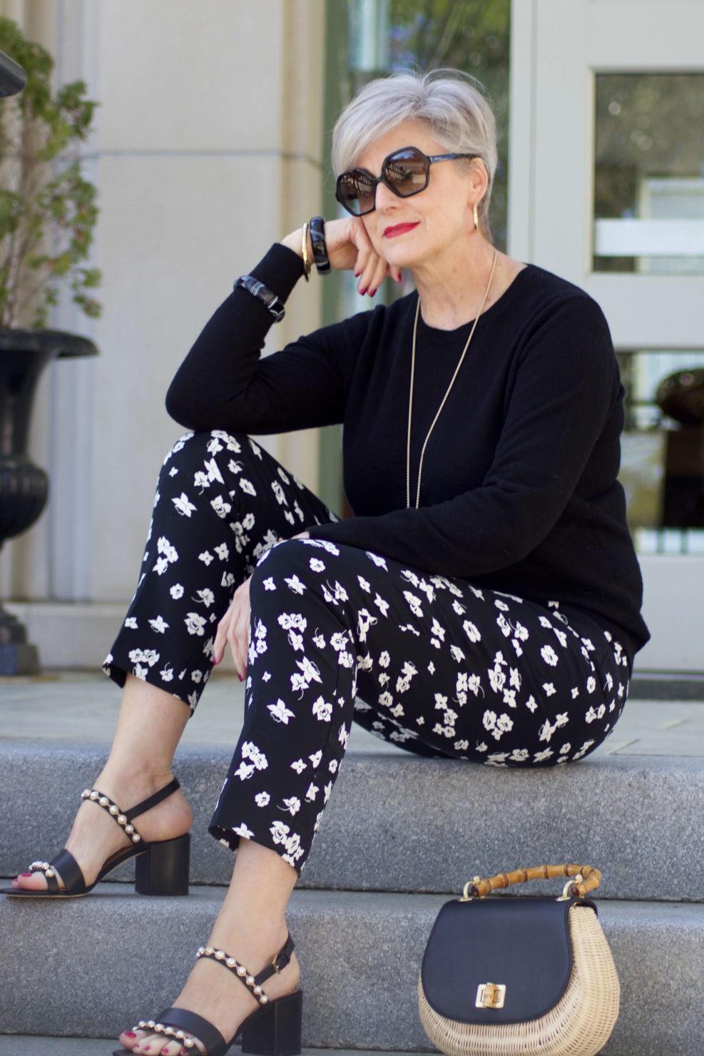 beth from Style at a Certain Age wears black and white floral ankle pants, black crewneck sweater, black pearl embellished sandals, and straw handbag