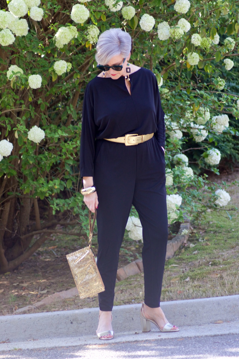 beth from Style at a Certain Age wears a black jumpsuit, gold sandals, gold belt, and gold clutch handbag