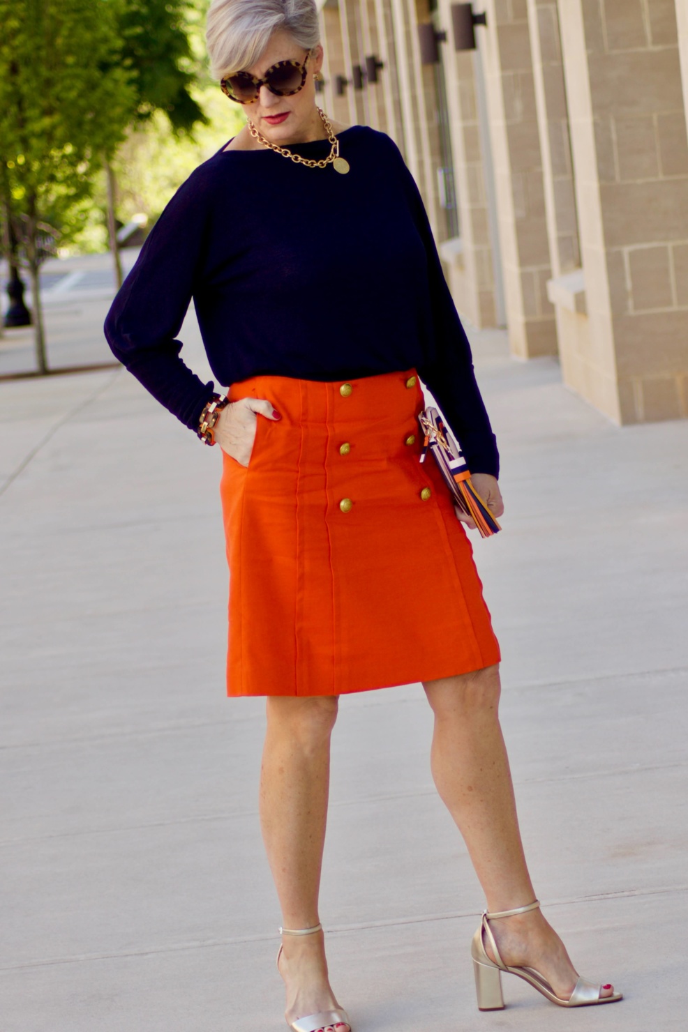 beth from Style at a Certain Age wears a J.Crew orange skirt, blue tee, gold sandals, and Tory Burch mini bag.