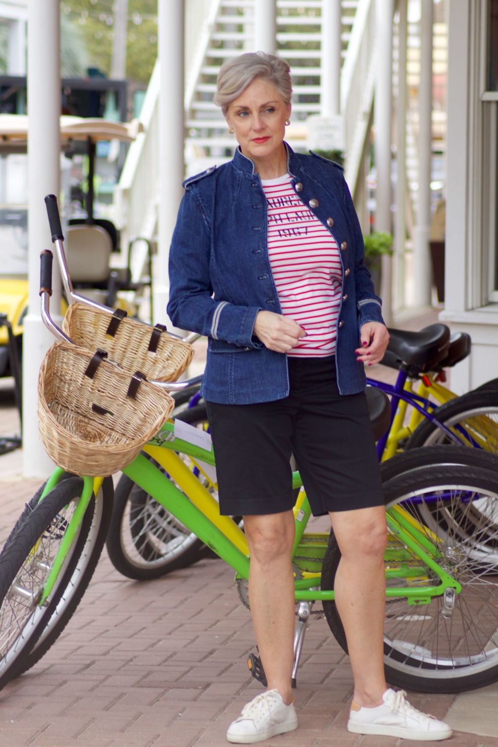 beth from Style at a Certain Age wears a military inspired denim jacket, red stripe tee, black bermuda shorts, and white tennis shoes