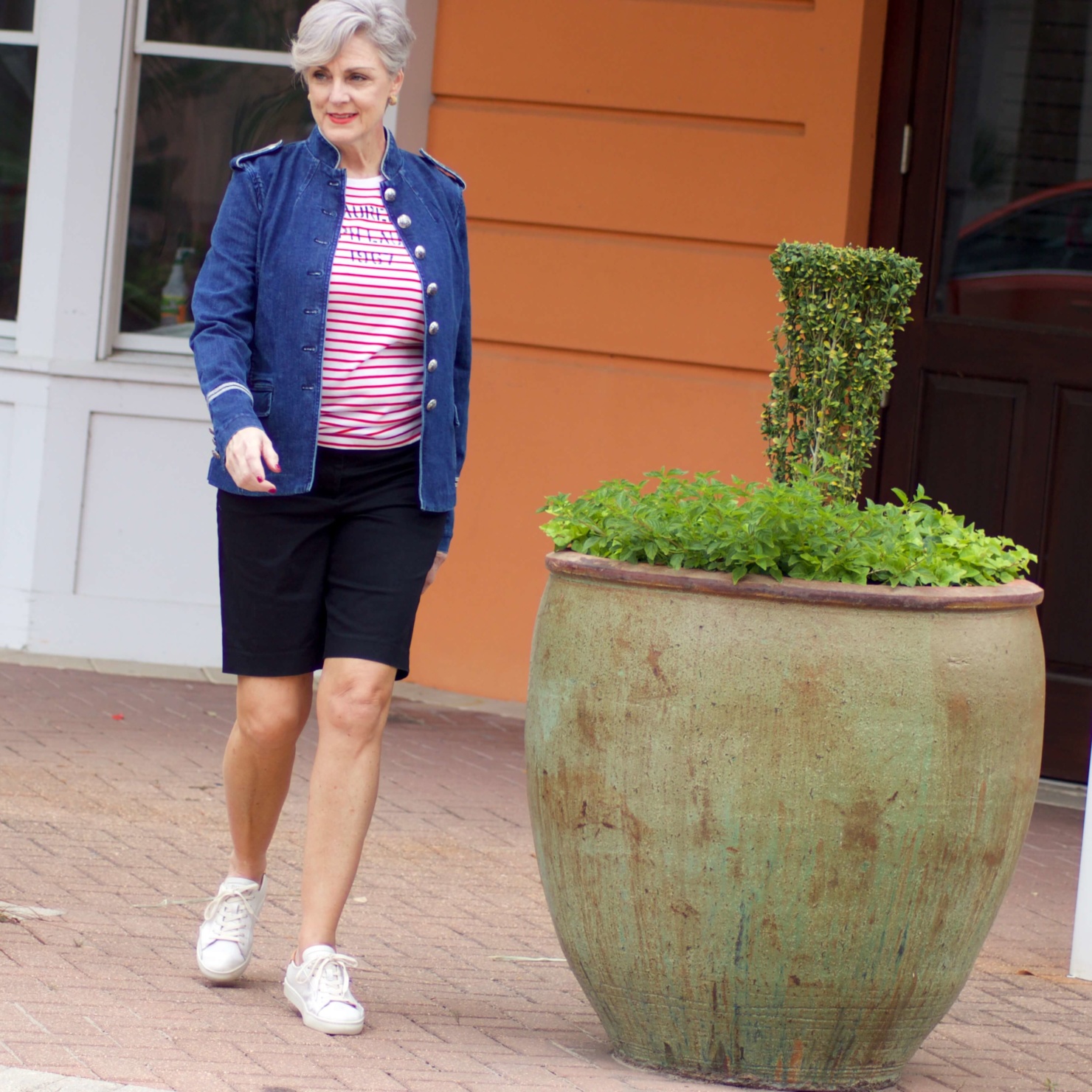 beth from Style at a Certain Age wears a military inspired denim jacket, red stripe tee, black bermuda shorts, and white tennis shoes