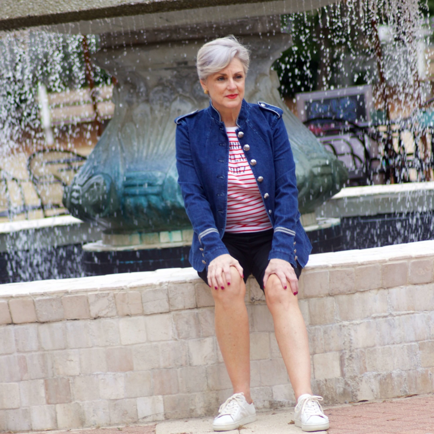 beth from Style at a Certain Age wears a military inspired denim jacket, red stripe tee, black bermuda shorts, and white tennis shoes