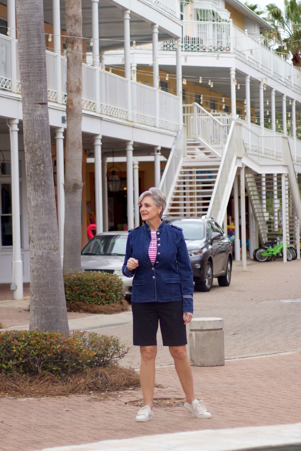 beth from Style at a Certain Age wears a military inspired denim jacket, red stripe tee, black bermuda shorts, and white tennis shoes