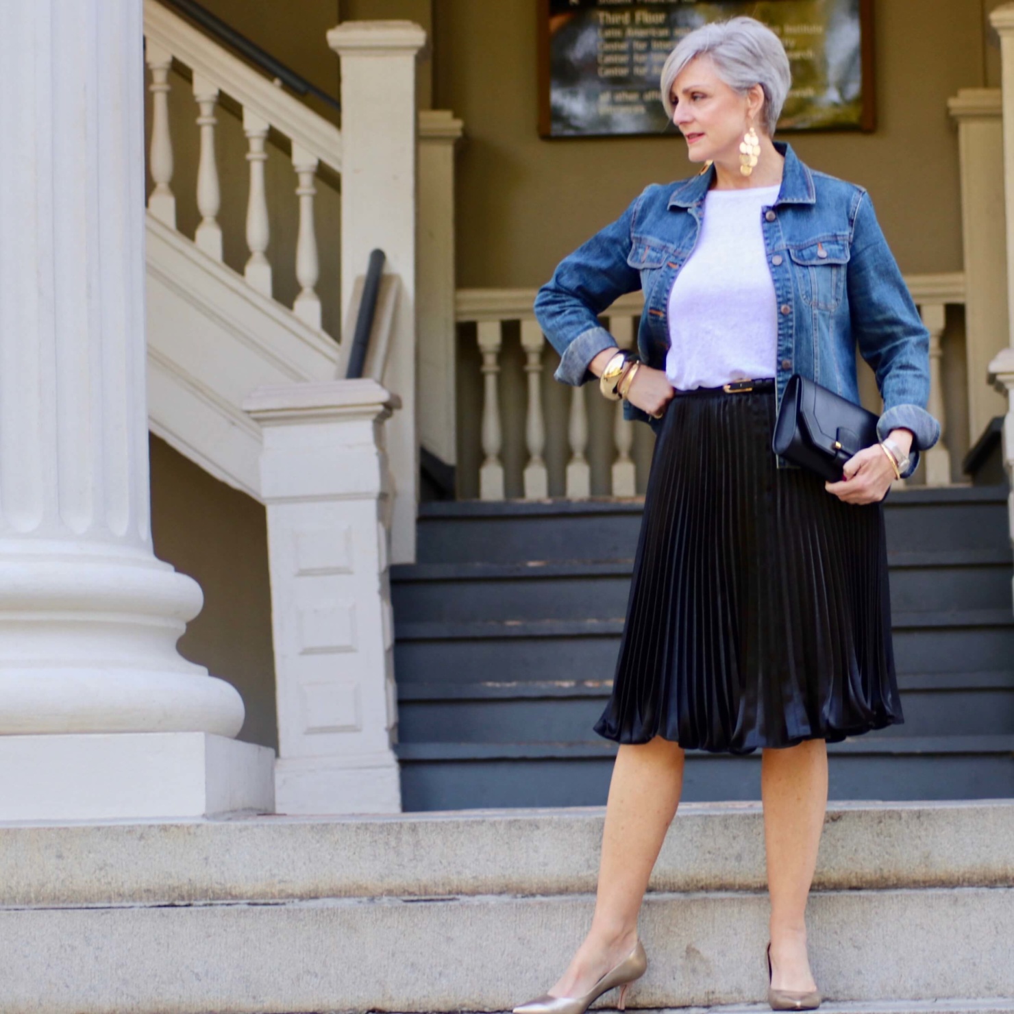 beth from Style at a Certain Age wears a black pleated skirt, white tee, denim jacket, and gold metallic pumps