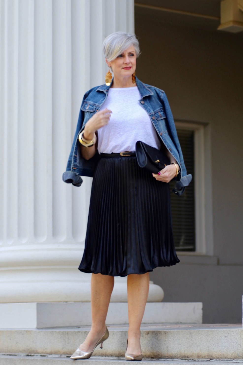 beth from Style at a Certain Age wears a black pleated skirt, white tee, denim jacket, and gold metallic pumps