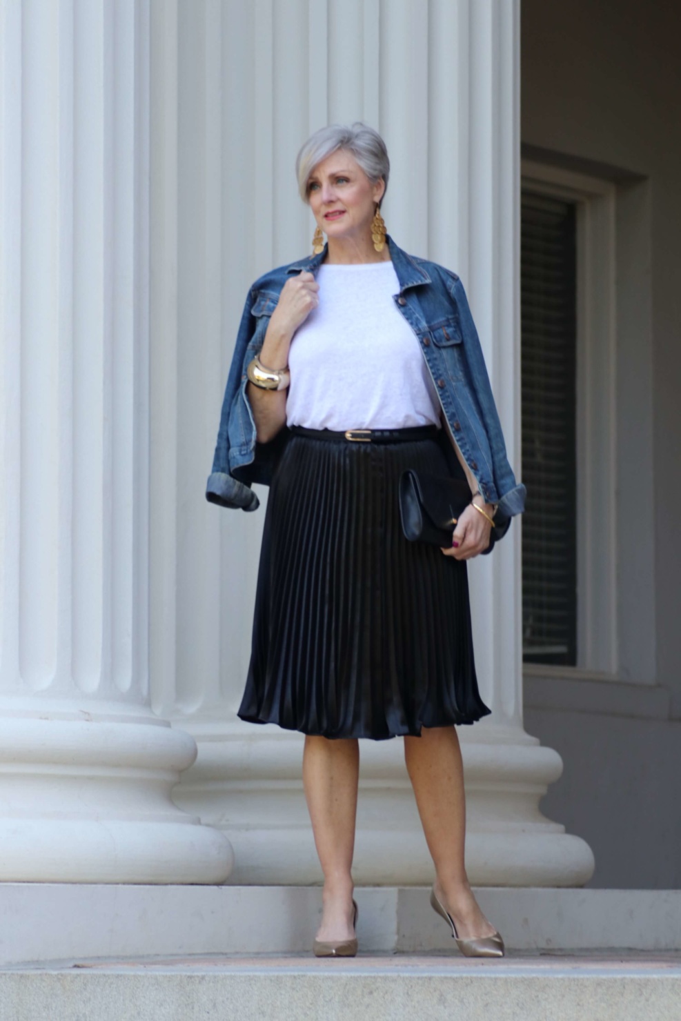 beth from Style at a Certain Age wears a black pleated skirt, white tee, denim jacket, and gold metallic pumps