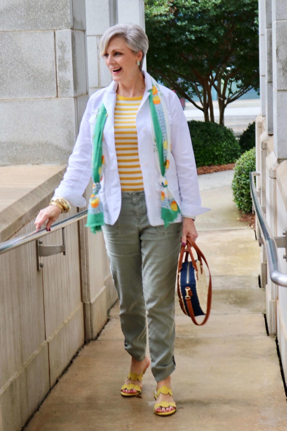 how to wear a jacket for spring and summer beth from Style at a Certain Age wears a white utility jacket, striped tee, green utility pants, yellow sandals, lemon scarf, and cornhusk handbag