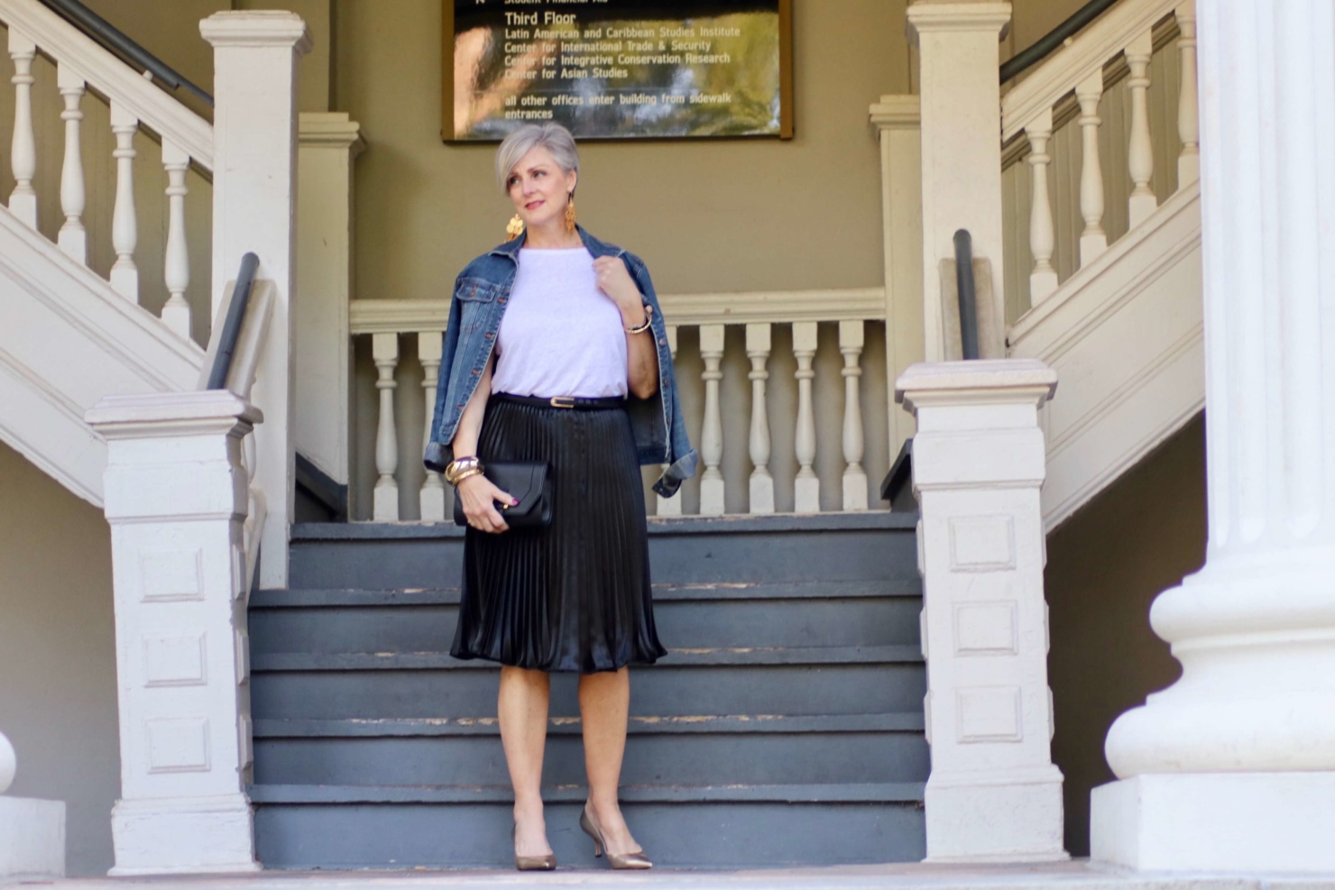 beth from Style at a Certain Age wears a black pleated skirt, white tee, denim jacket, and gold metallic pumps