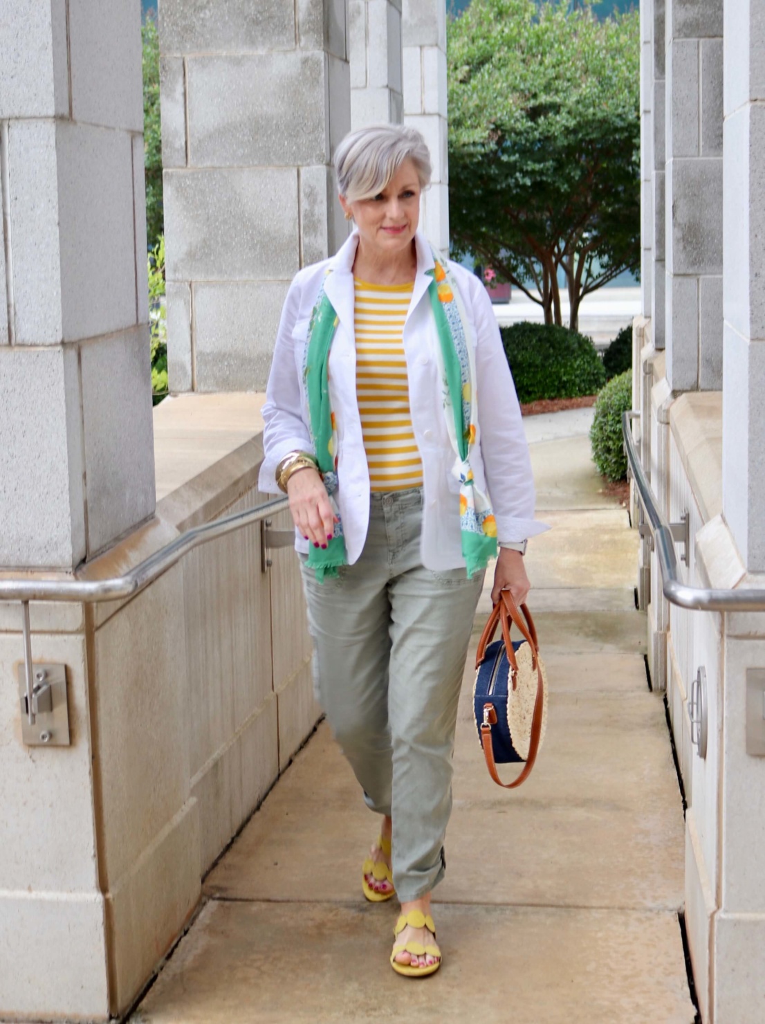 how to wear a jacket for spring and summer beth from Style at a Certain Age wears a white utility jacket, striped tee, green utility pants, yellow sandals, lemon scarf, and cornhusk handbag