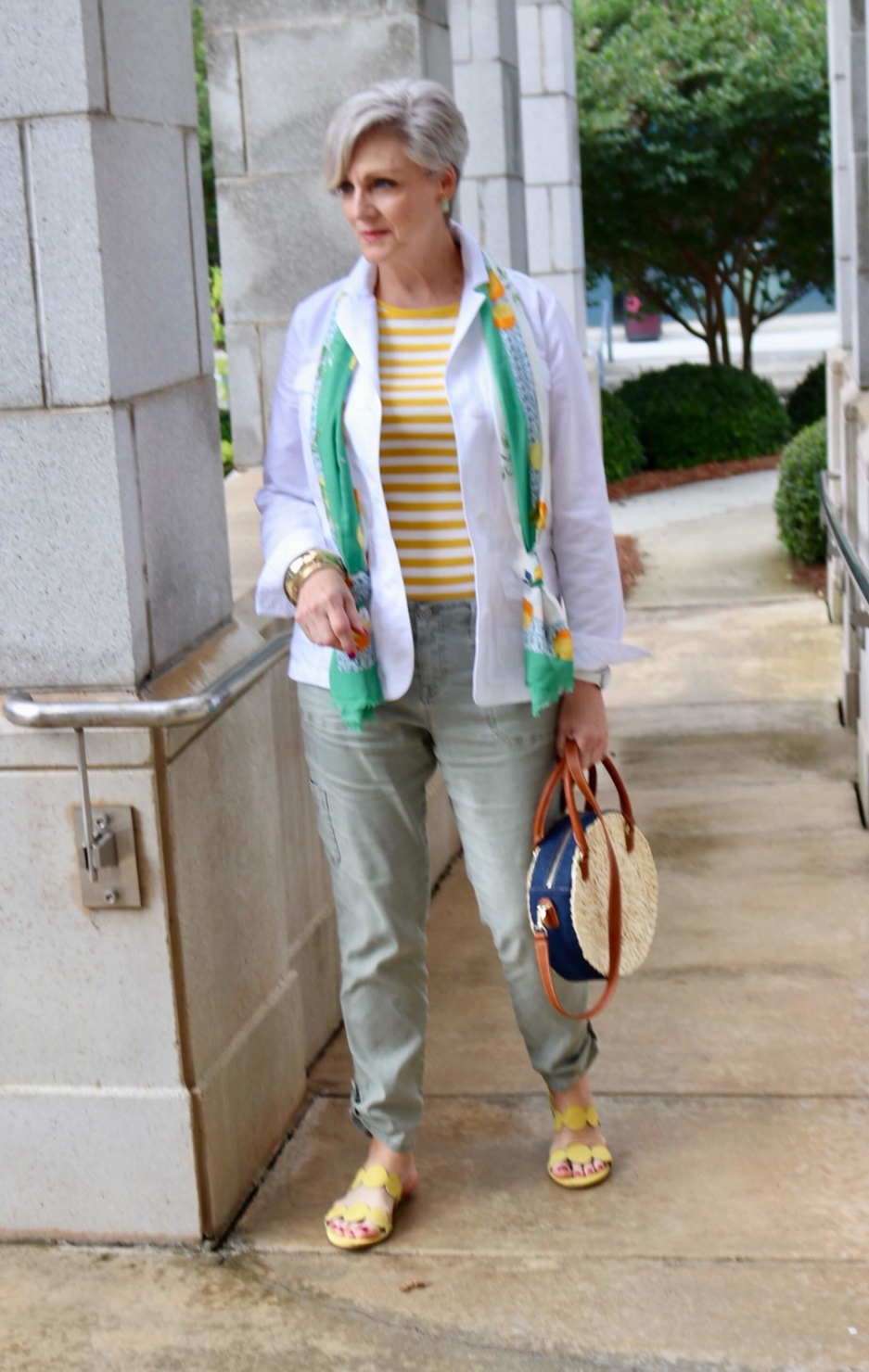 how to wear a jacket for spring and summer beth from Style at a Certain Age wears a white utility jacket, striped tee, green utility pants, yellow sandals, lemon scarf, and cornhusk handbag