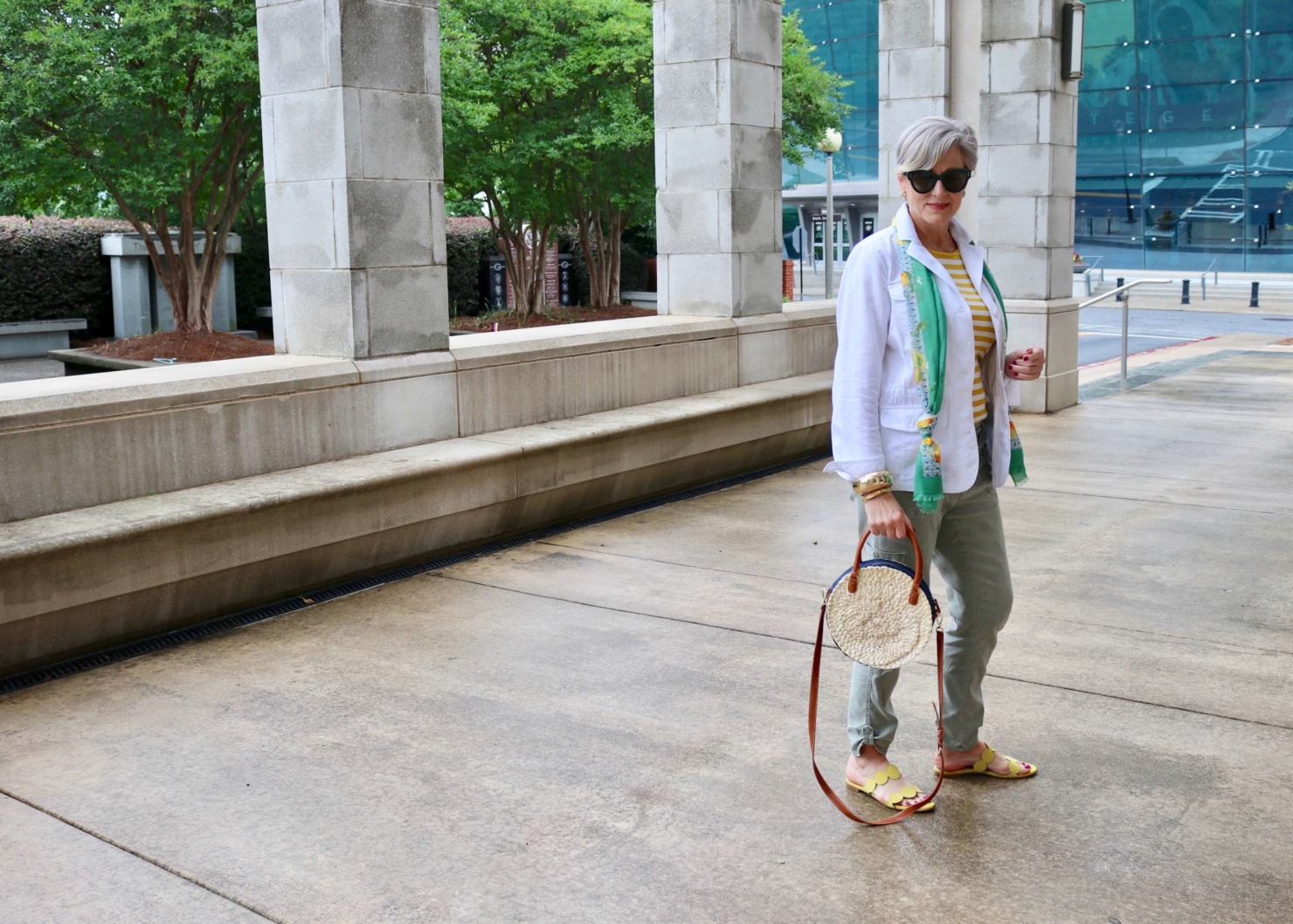 how to wear a jacket for spring and summer beth from Style at a Certain Age wears a white utility jacket, striped tee, green utility pants, yellow sandals, lemon scarf, and cornhusk handbag