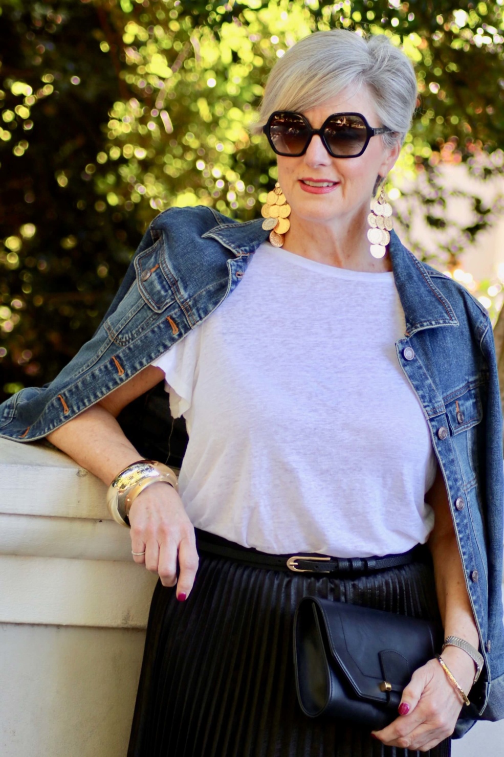 beth from Style at a Certain Age wears a black pleated skirt, white tee, denim jacket, and gold metallic pumps