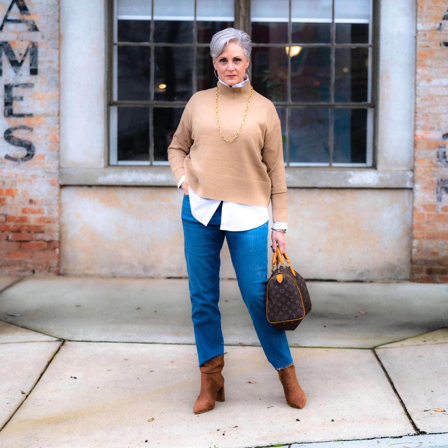 Woman over 50 wearing a camel knit over a white shirt with blue jeans and brown ankle boots.