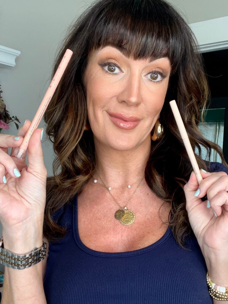 Jennifer smiling at the camera showing two light-colored eye pencils for lining the water line 