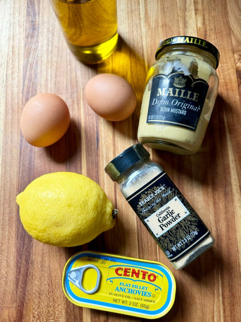 Flat lay of Caesar-style dressing ingredients on a wood cutting board: two brown eggs, a jar of Maille Dijon mustard, Trader Joe's garlic powder, a lemon, a bottle of olive oil, and a can of Cento flat fillet anchovies