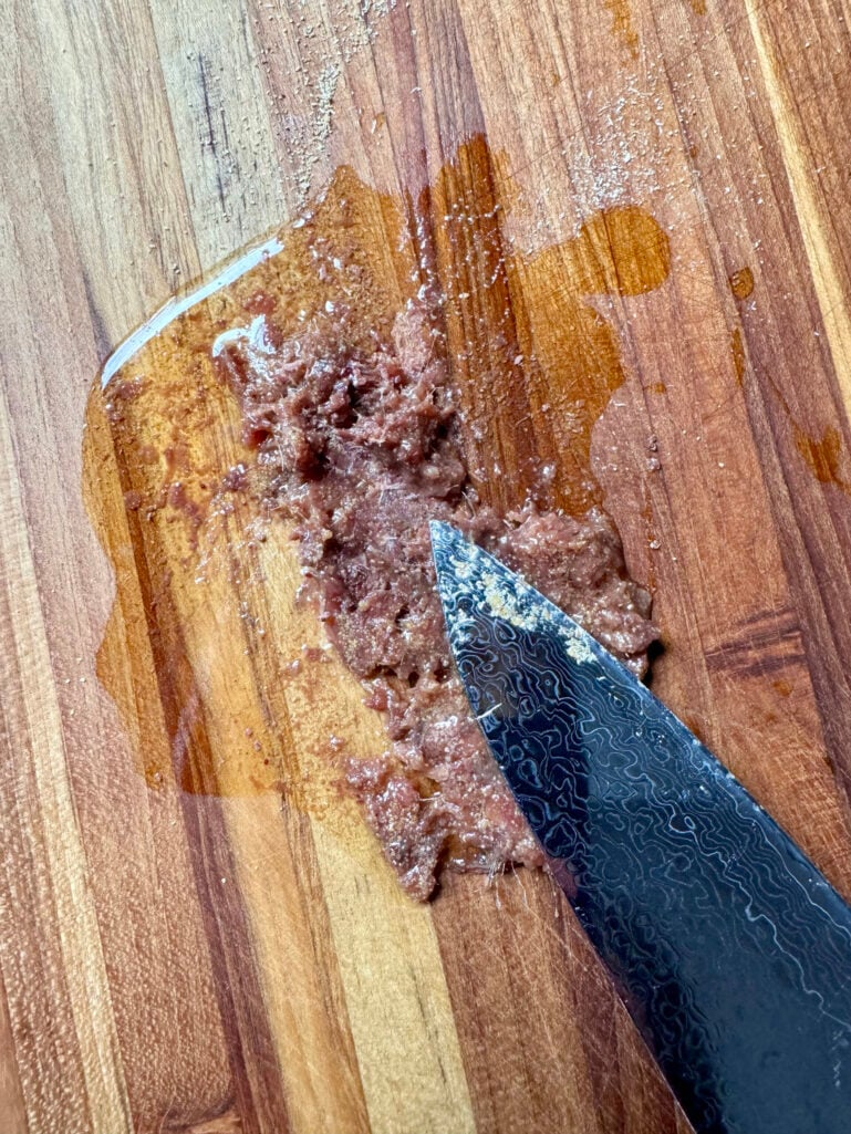 Anchovy fillets being smashed into a paste with the flat side of a Damascus steel chef's knife on a wood cutting board