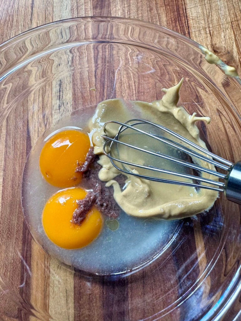 Two bright orange egg yolks, Dijon mustard, and anchovy paste in a glass mixing bowl with a whisk