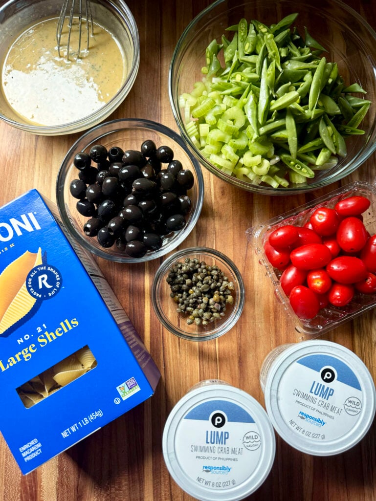 Overhead flat lay of salad ingredients on a wood cutting board: finished Caesar dressing, sliced snap peas and celery, black olives, capers, grape tomatoes, two tubs of Publix lump crab meat, and a box of Ronzoni large shell pasta