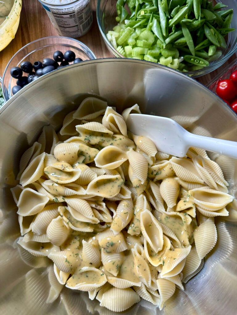 Large pasta shells tossed with Caesar-style anchovy dressing in a stainless mixing bowl with a white spatula, with salad ingredients visible in the background